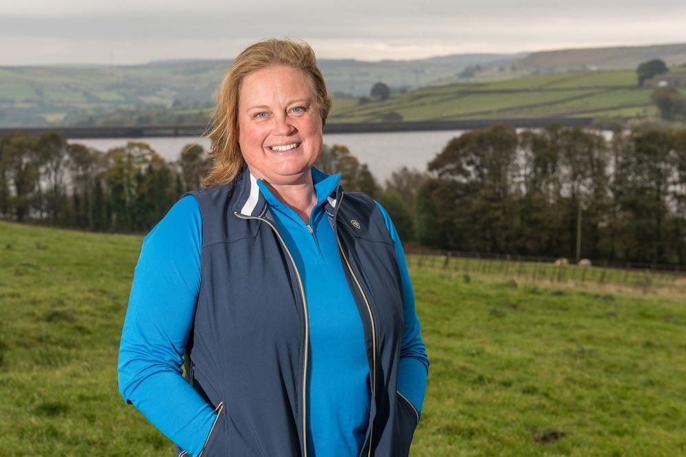A farmer stands on her farm with a beautiful backdrop behind her.