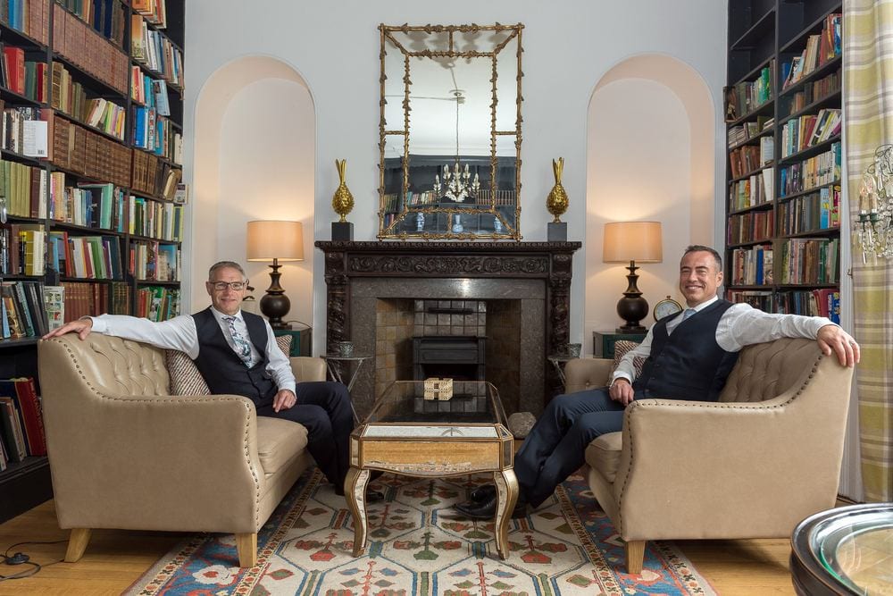 Two men in matching waistcoats, seated either side of a library fireplace.
