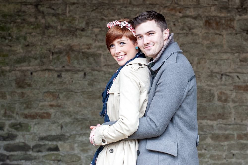 A young couple cuddle in front of an old brick wall.